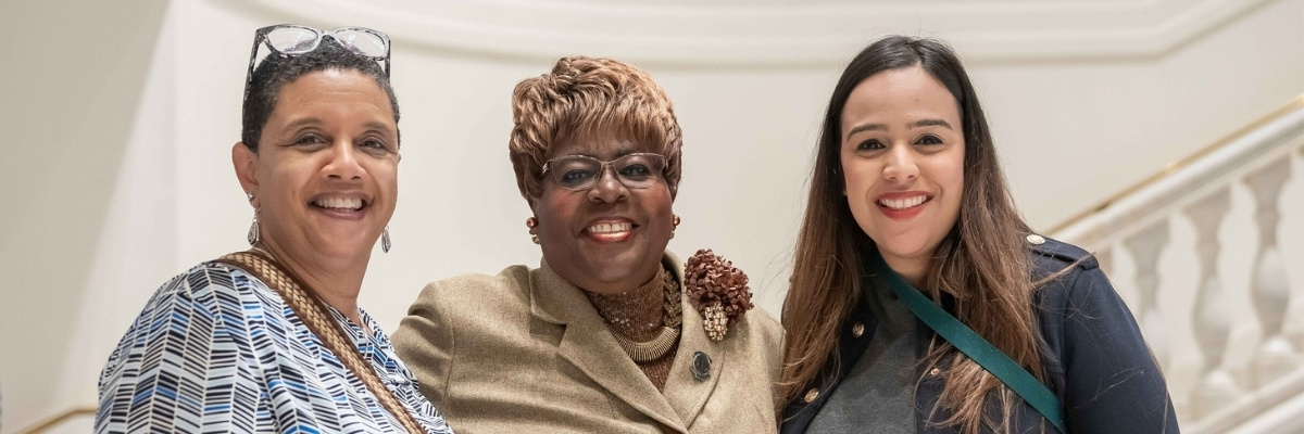 Three women are standing next to each other in a bright room in front of a staircase, posing for a group shot. They have light, medium-light and dark skin tones.